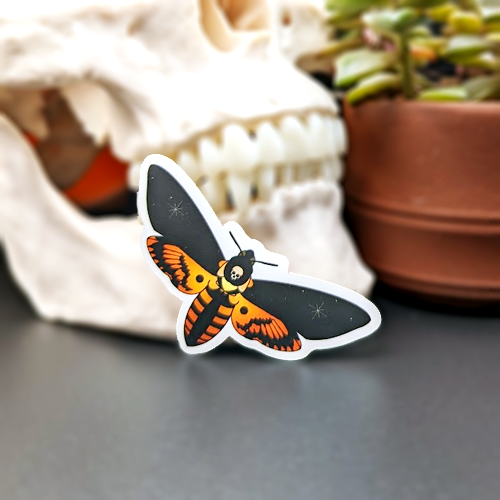 A sticker depicting a death's-head hawkmoth on a reflective surface with a blurred background